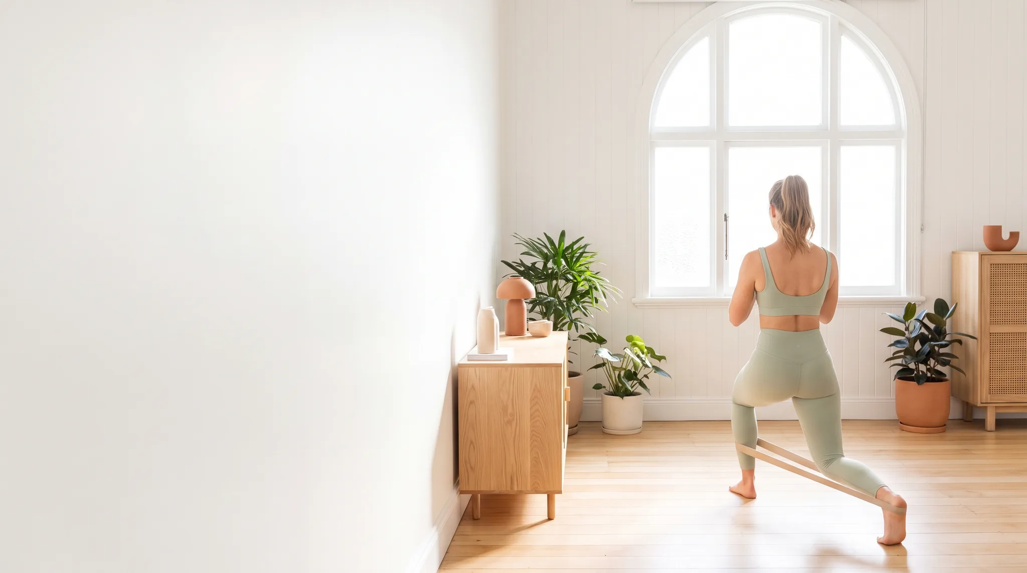 A woman does a resistance band lunge with a physiotherapist coaching her form in a bright, plant-filled studio